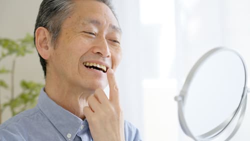 年長男士對著鏡子檢查牙齒,微笑露出活動假牙,象徵假牙佩戴後的生活自信。